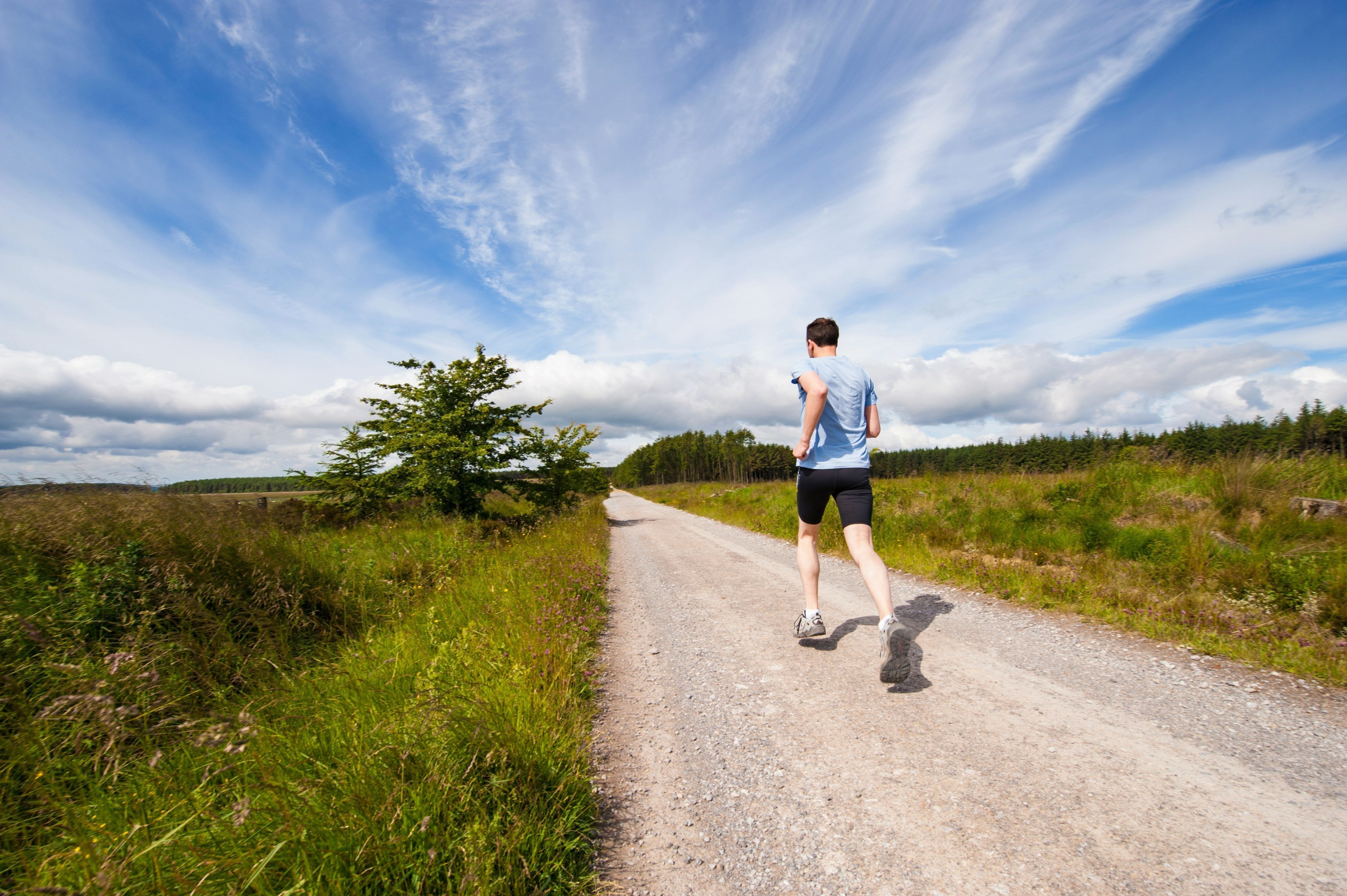 A man running down a gravel track with a beautiful blue and cloudy skyline, and bushes, trees and grass on the verges