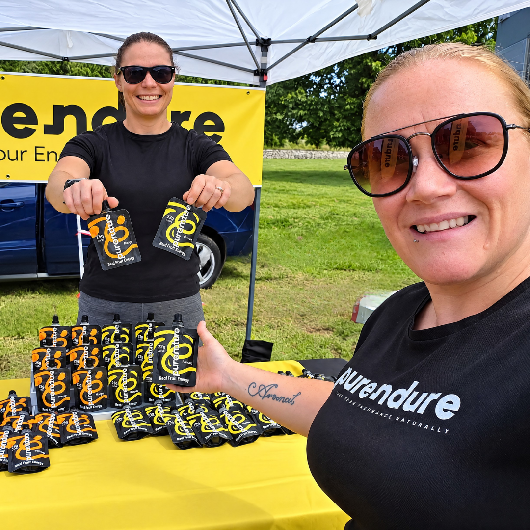 Two women at an outdoor booth under a white canopy, holding and displaying black and yellow pouches branded Aurendure. A yellow banner and table are visible in the background, with grass and trees outside.