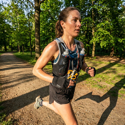 Woman running on a trail with a hydration vest in a forest setting holding a Purendure pouch